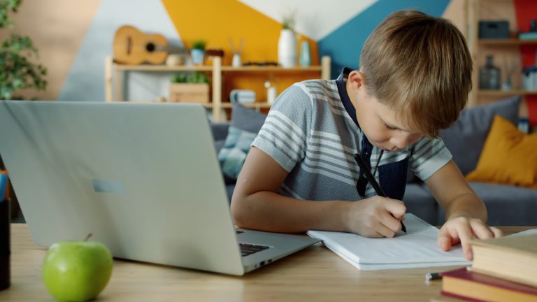 Child writing in a notebook beside a laptop at home